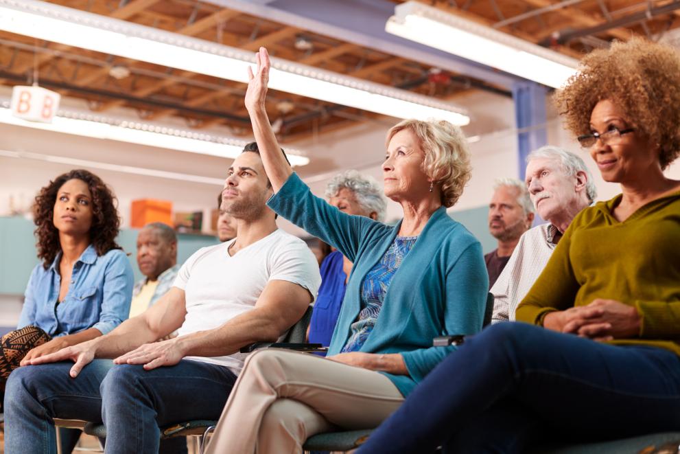 A woman in the front row of a neighbourhood meeting in a community centre with her hands raised.