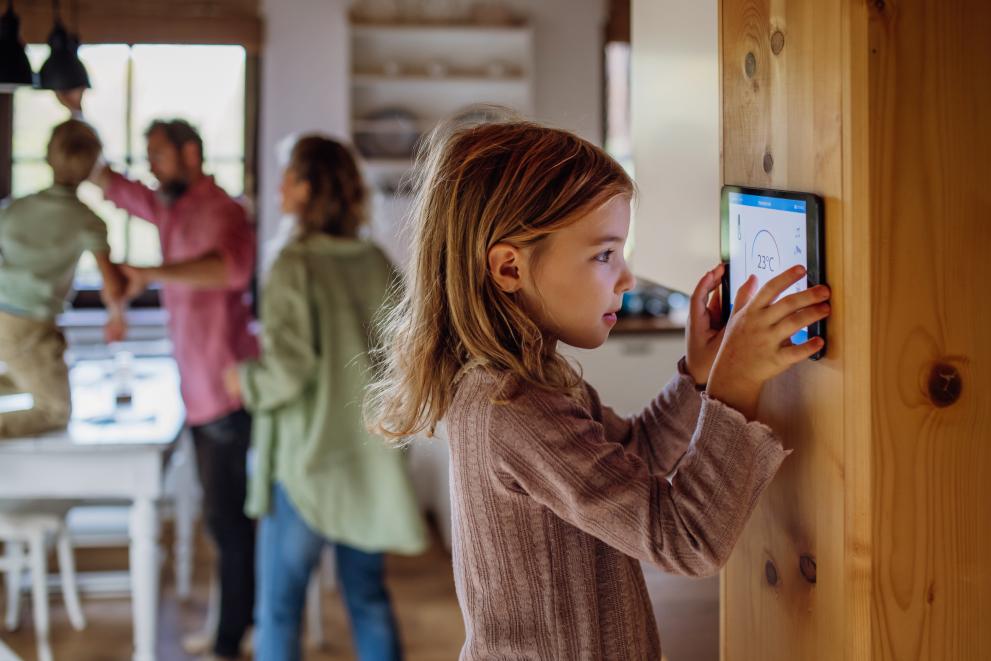 A young girl is looking at a smart energy monitor in her dining room, while in the background her parents and brother are focused on fitting a lightbulb.