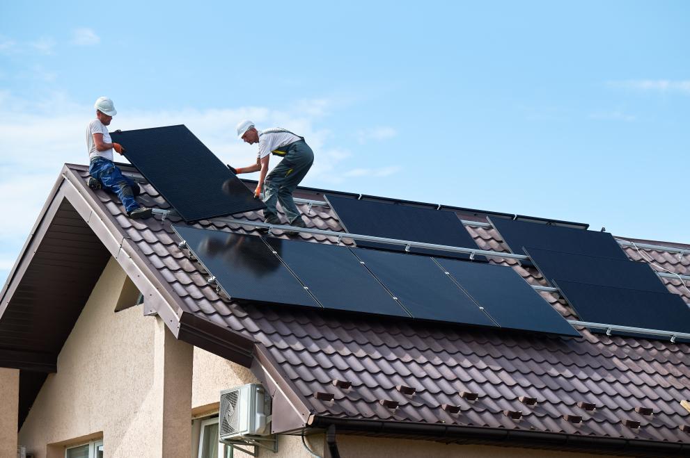 Two male installers in helmets carrying a photovoltaic solar module, building a solar panel system on the roof of a house.