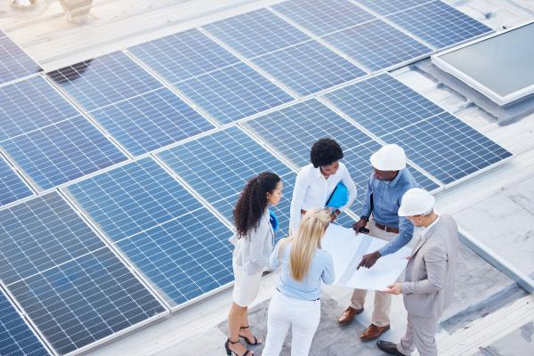 Two engineering consultants in hard hats are standing on a roof, next to solar panels, with three female businesspeople. They are explaining solar energy designs to them.