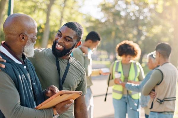 A scene in a park shows a smiling man wearing a lanyard, who has his arm around an older man who is showing him a tablet. In the background are three people also looking at tablets, one is wearing a high vis.