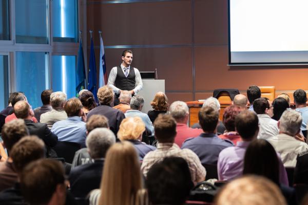 A speaker talking during a citizen energy panel in a conference hall. Rear view of diverse citizens in the audience.