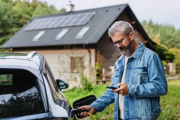 A man charging an electric car in front of his house, plugging the charger into the charging port. A house with a solar panel system on the roof behind him.